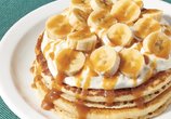 La imagen muestra los NUEVOS pancakes de plátano y caramelo salado: dos pancakes cubiertos con rodajas de plátano, crema de vainilla rociados con caramelo.