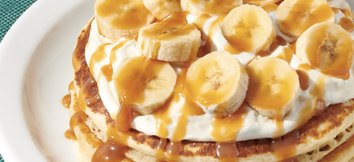 La imagen muestra los NUEVOS pancakes de plátano y caramelo salado: dos pancakes cubiertos con rodajas de plátano, crema de vainilla rociados con caramelo.