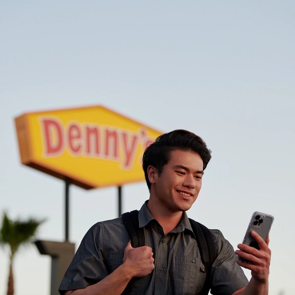 Hombre con un teléfono al aire libre, con el cartel de Denny's de fondo