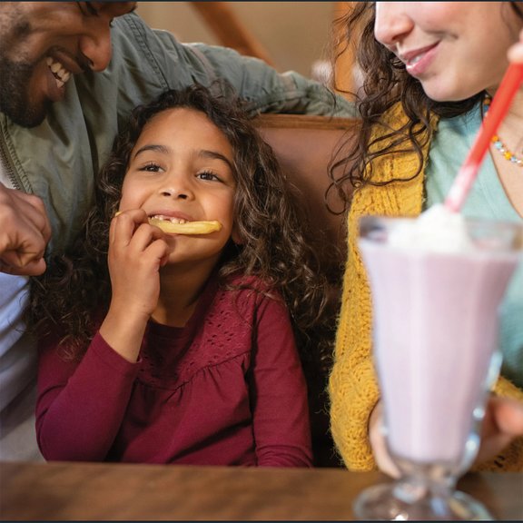 Denny's Kids Eat Free image with young girl eating a french fry & milkshake